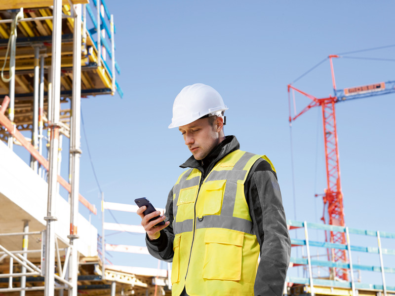 Eine Person mit Sicherheitsausrüstung überprüft auf einer Baustelle ein Smartphone. Anwendungsbild