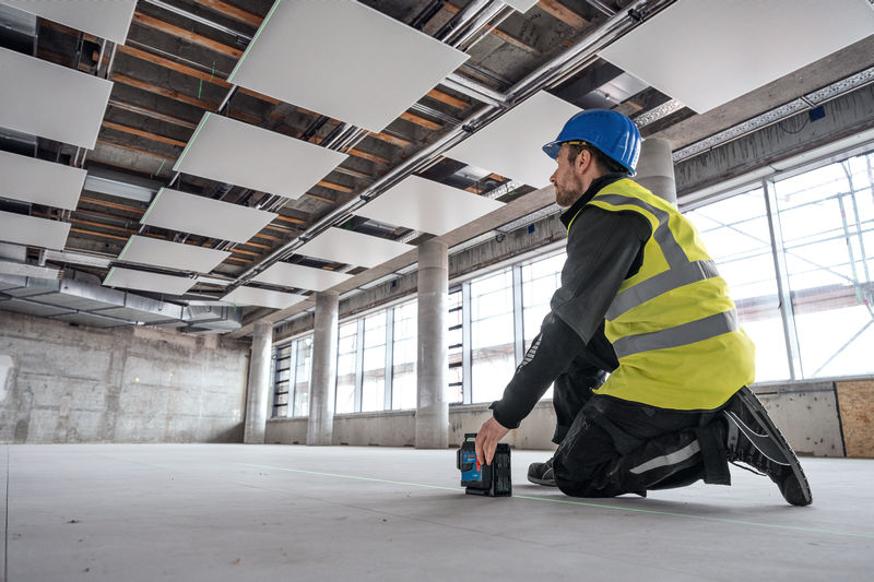 Eine Person mit Sicherheitsausrüstung verwendet ein Lasernivelliergerät auf dem Boden einer großen Baustelle. Anwendungsbild