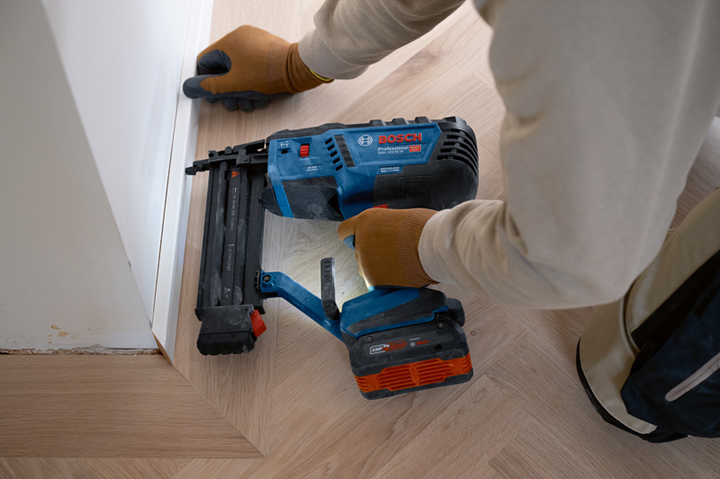 Eine Person mit Sicherheitsausrüstung befestigt mit einem Akku-Holznagler Zierleisten am Boden. Anwendungsbild