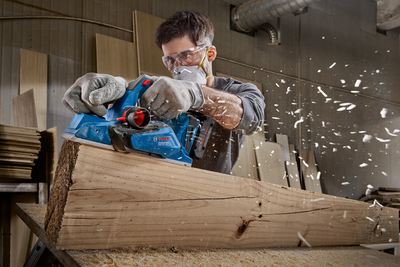 Eine Person mit Schutzausrüstung glättet einen großen Holzbalken mit einem Elektrohobel. Anwendungsbild