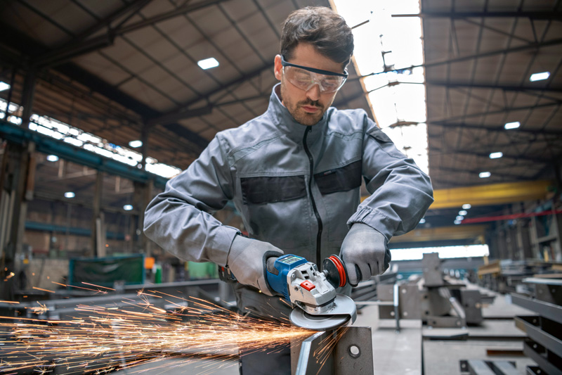 Eine Person mit Schutzausrüstung schleift in einer Industriewerkstatt Metall und erzeugt dabei Funken. Anwendungsbild