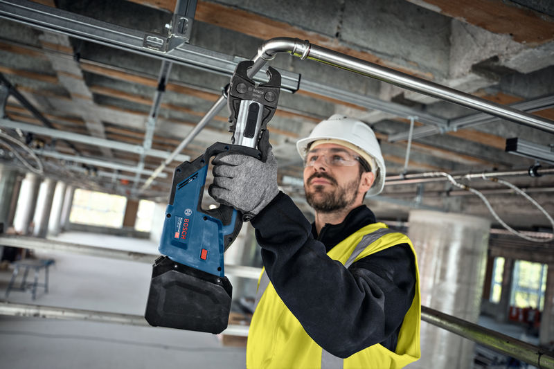 Eine Person mit Schutzausrüstung verwendet ein Akku-Presswerkzeug an einem Metallrohr. Anwendungsbild