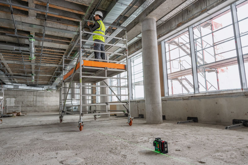 Eine Person mit Sicherheitsausrüstung arbeitet auf einem Gerüst und projiziert mit einem Lasernivelliergerät Linien. Anwendungsbild