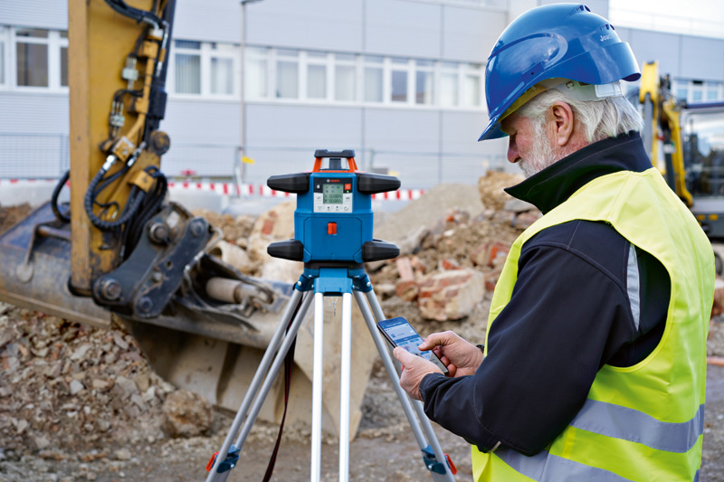 Eine Person mit Sicherheitsausrüstung bedient auf einer Baustelle ein Lasernivelliergerät. Anwendungsbild