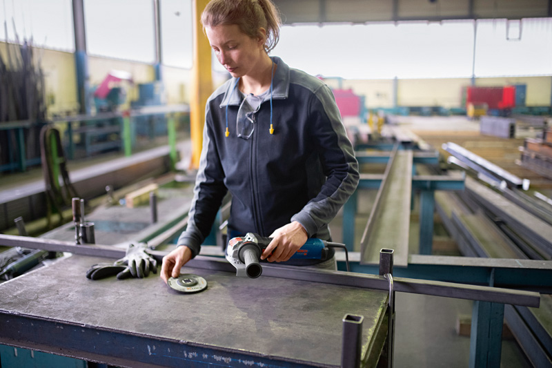 Eine Person mit Schutzausrüstung bereitet einen Winkelschleifer auf einer Metallwerkbank vor. Anwendungsbild