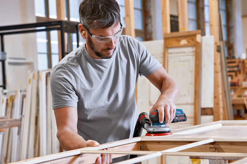 Eine Person mit Schutzausrüstung schleift in einer Werkstatt Holz mit einem elektrischen Schleifgerät. Anwendungsbild