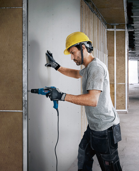 Eine Person mit Sicherheitsausrüstung bohrt in die Trockenbauwand eines im Bau befindlichen Gebäudes. Anwendungsbild