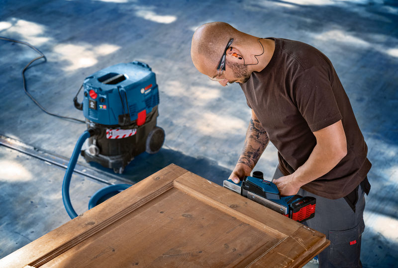 Eine Person mit Schutzausrüstung schleift eine Holztür mit einem Elektrohobel. Anwendungsbild