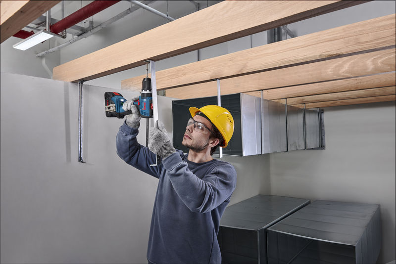 Eine Person mit Schutzausrüstung bohrt in eine Metallstange unter Holzbalken. Anwendungsbild