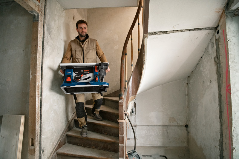 Eine Person trägt eine kabellose Tischkreissäge eine schmale Treppe auf einer Baustelle hinunter. Anwendungsbild