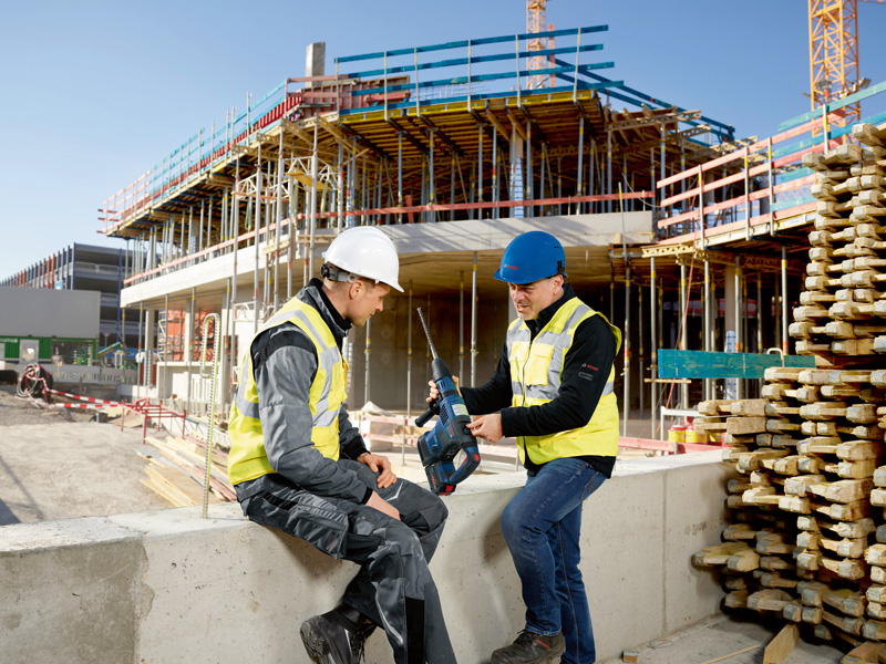 Zwei Personen in Sicherheitsausrüstung besprechen auf einer Baustelle einen Bohrhammer. Anwendungsbild
