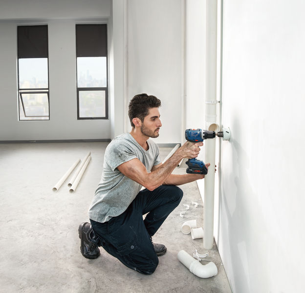 Eine Person bohrt in eine Wand neben Wasserrohren in einem teilweise fertiggestellten Raum. Anwendungsbild