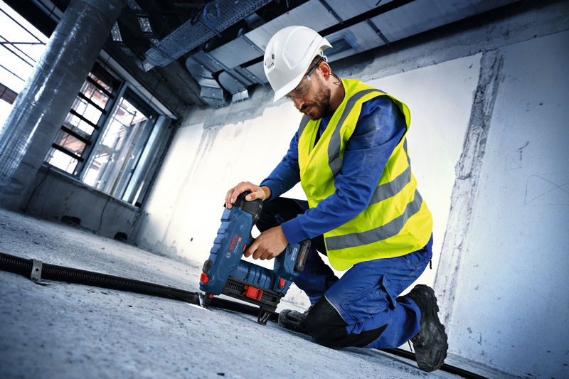Eine Person mit Sicherheitsausrüstung bohrt mit einem Elektrowerkzeug in einen Betonboden. Anwendungsbild