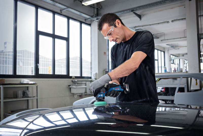 Eine Person mit Sicherheitsausrüstung poliert in einer Autowerkstatt eine Motorhaube. Anwendungsbild