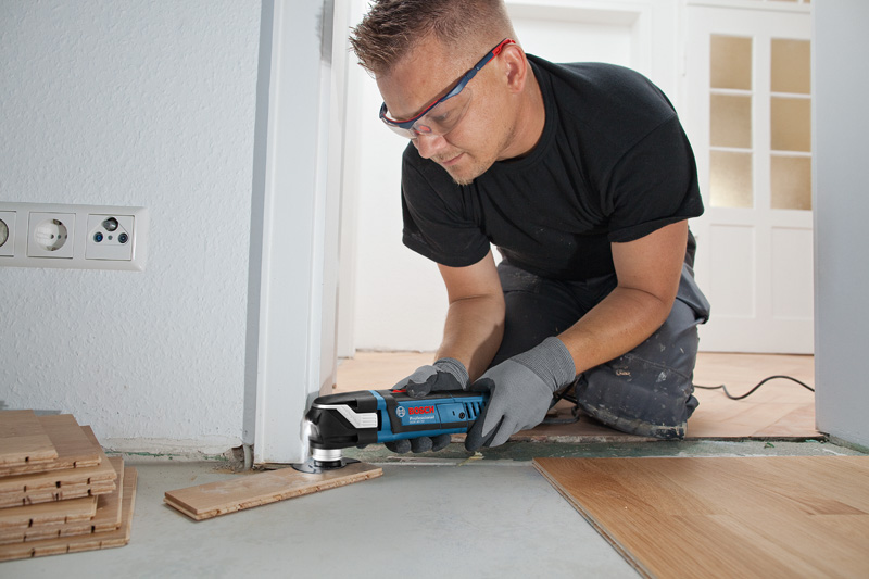 Eine Person mit Sicherheitsausrüstung schneidet mit einem Multischneider in der Nähe einer Tür Dielenbretter zu. Anwendungsbild