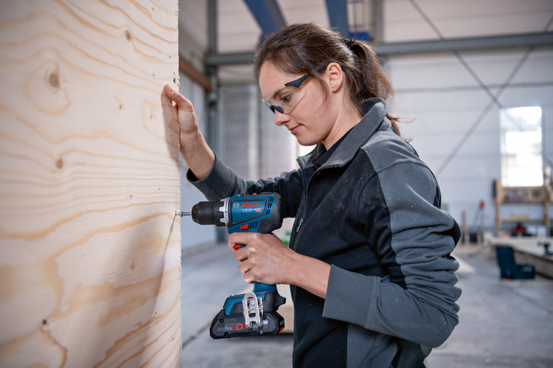 Eine Person mit Schutzausrüstung bohrt in einer Werkstatt in ein Holzbrett. Anwendungsbild