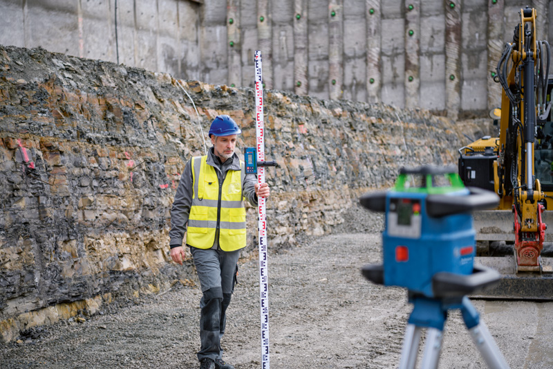 Eine Person mit Sicherheitsausrüstung verwendet auf einer Baustelle einen Laserempfänger mit einem Nivelliergerät. Anwendungsbild