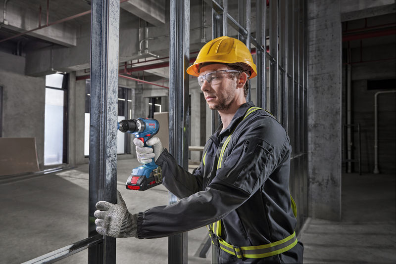 Eine Person mit Sicherheitsausrüstung bohrt auf einer Baustelle in ein Metallgerüst. Anwendungsbild