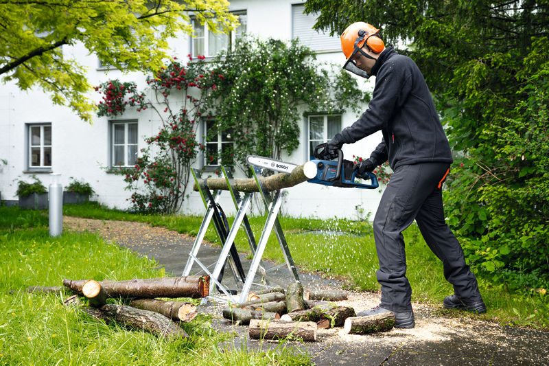 Eine Person mit Sicherheitsausrüstung sägt im Freien mit einer Akku-Kettensäge Holzstämme. Anwendungsbild