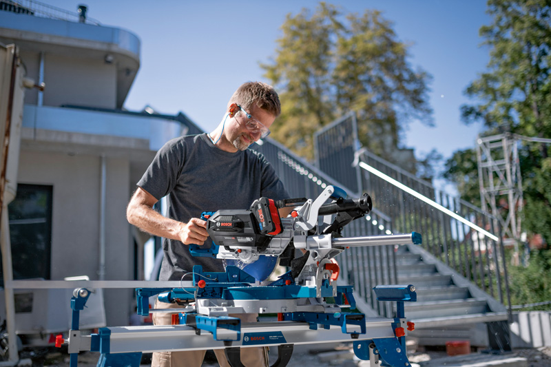 Eine Person mit Sicherheitsausrüstung bedient auf einer Baustelle eine kabellose Gehrungssäge. Anwendungsbild