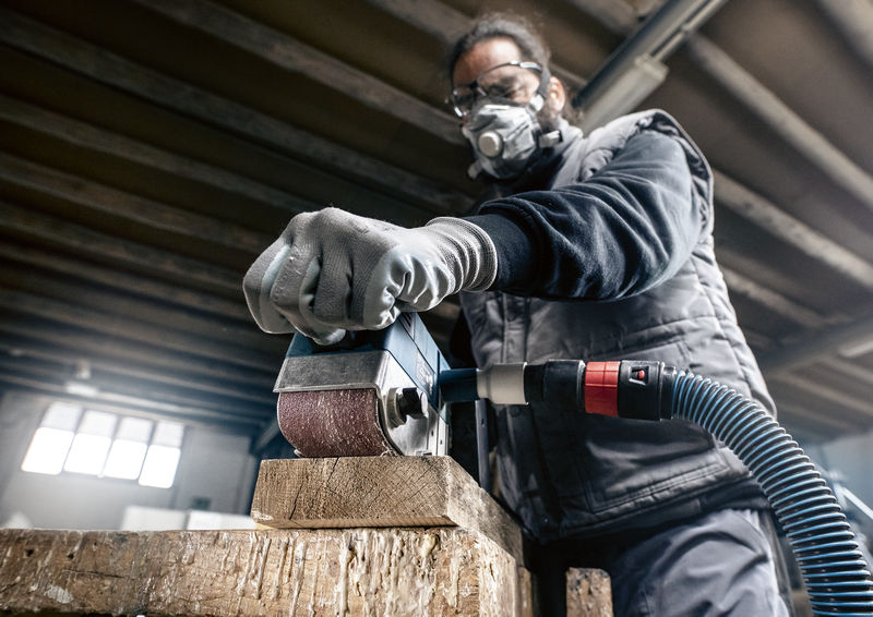 Eine Person mit Schutzausrüstung schleift Holz mit einem staubabsaugenden Elektrowerkzeug. Anwendungsbild