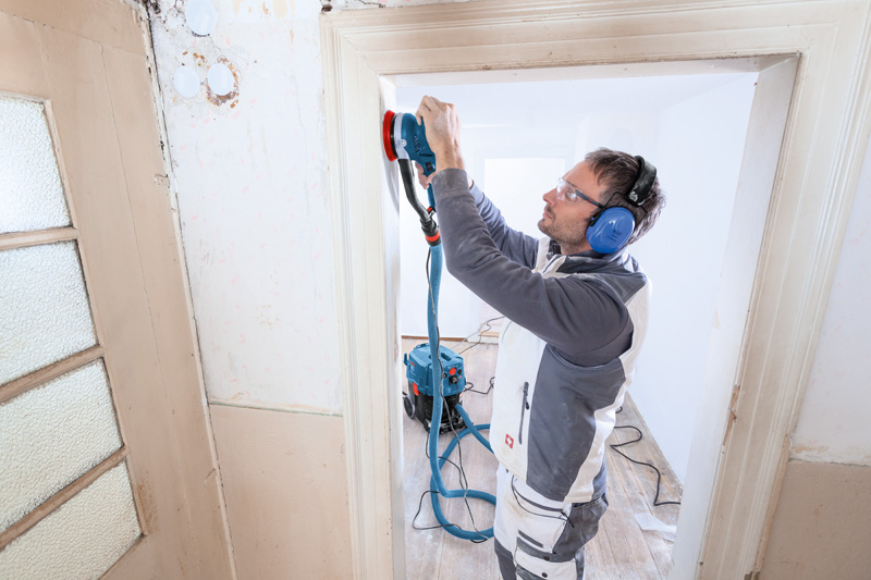 Eine Person mit Schutzausrüstung schleift einen Türrahmen mit einem Exzenterschleifer. Anwendungsbild