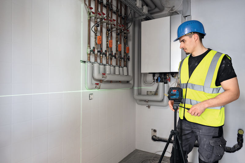 Eine Person mit Sicherheitsausrüstung bedient in einem Hauswirtschaftsraum ein Lasernivelliergerät. Anwendungsbild