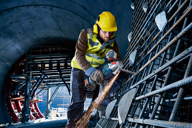 Ein Arbeiter mit Sicherheitsausrüstung schleift in einem Tunnel Bewehrungsstahl mit einem Akku-Winkelschleifer. Anwendungsbild