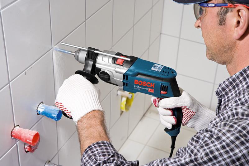 Eine Person mit Schutzausrüstung bohrt mit einer Schlagbohrmaschine in eine geflieste Wand. Anwendungsbild