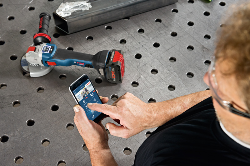 Eine Person mit Schutzausrüstung steuert mit einem Smartphone einen Akku-Schleifapparat auf einer Metallwerkbank. Anwendungsbild