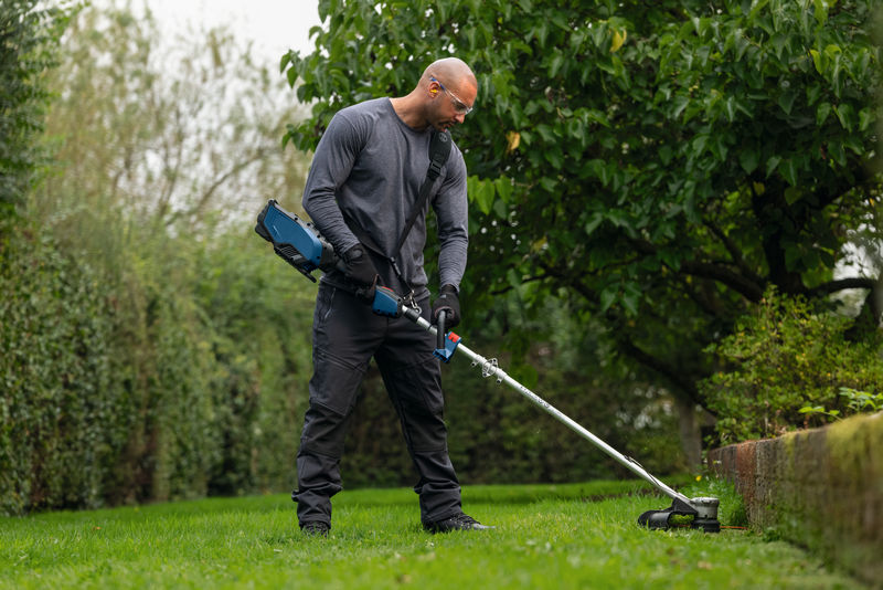 Eine Person mit Sicherheitsausrüstung schneidet mit einem Akku-Gerät das Gras entlang einer Gartenkante. Anwendungsbild