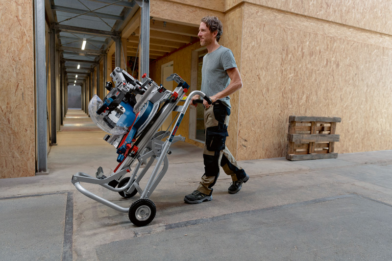 Eine Person mit Sicherheitsausrüstung bewegt eine Akku-Gehrungssäge auf einem Rollständer im Innenbereich. Anwendungsbild