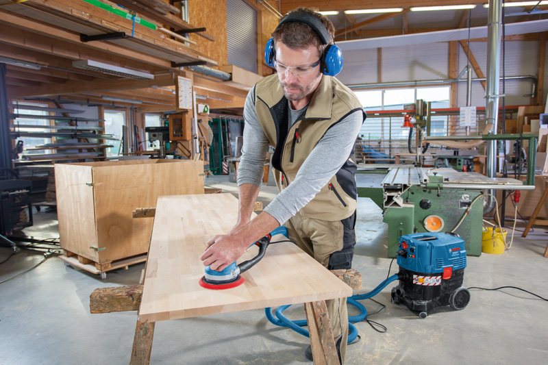 Person mit Schutzausrüstung schleift eine Holztischplatte mit einem Exzenterschleifer. Anwendungsbild