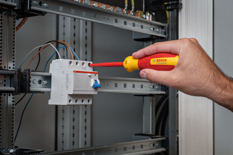 Eine Person zieht eine Schraube an einem elektrischen Bauteil in einem Bedienfeld fest. Anwendungsbild