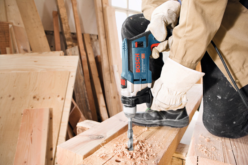Eine Person mit Schutzausrüstung bohrt in einer Werkstatt in ein Holzbrett. Anwendungsbild