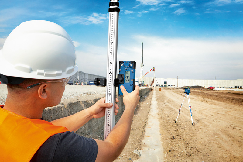 Eine Person mit Sicherheitsausrüstung verwendet auf einer Baustelle einen Laserempfänger mit einer Nivellierlatte. Anwendungsbild