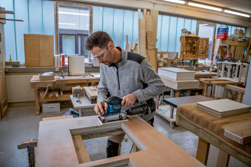 Eine Person mit Schutzausrüstung schleift in einer Werkstatt eine Holzplatte mit einem Schwingschleifer. Anwendungsbild