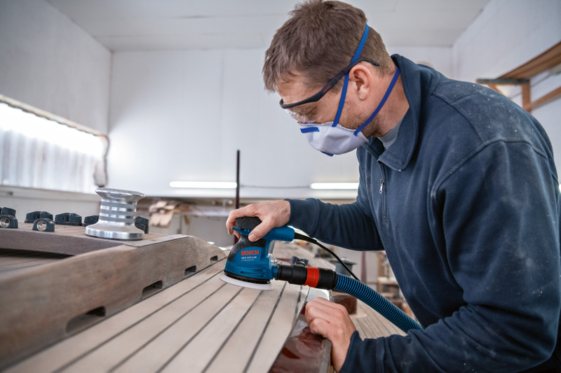 Eine Person mit Schutzausrüstung schleift Holzbretter mit einem Exzenterschleifer. Anwendungsbild
