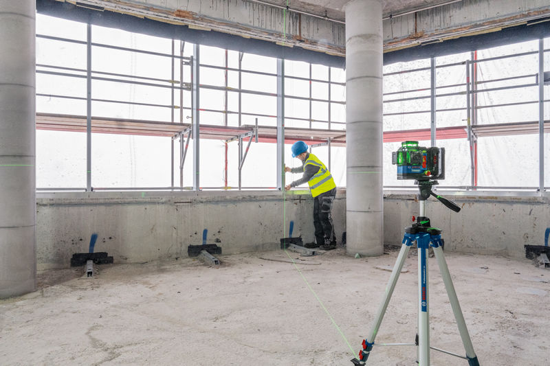 Eine Person mit Sicherheitsausrüstung richtet Materialien in der Nähe eines Lasernivelliergeräts auf einem Stativ aus. Anwendungsbild