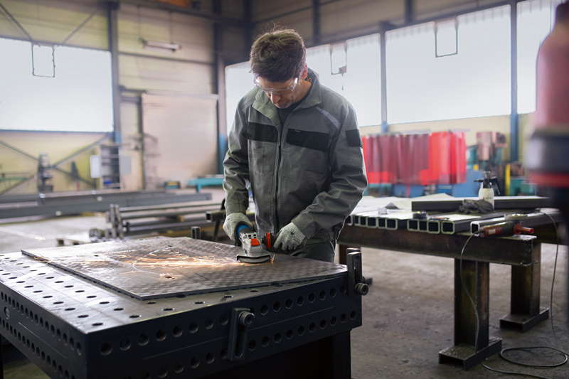 Ein Arbeiter mit Sicherheitsausrüstung schleift Metall auf einer großen Werkbank in einer Fabrik. Anwendungsbild