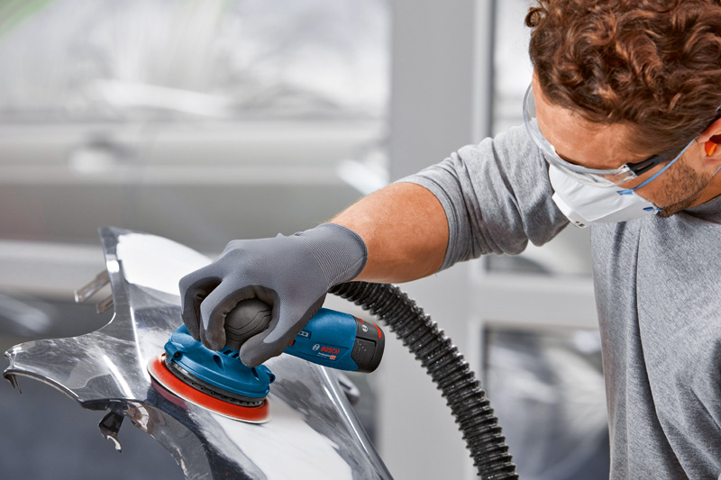 Eine Person mit Schutzausrüstung schleift ein Autoteil mit einem Akku-Exzenterschleifer. Anwendungsbild