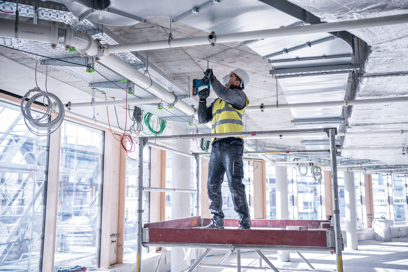 Eine Person mit Sicherheitsausrüstung bohrt auf einer Baustelle in eine Betondecke. Anwendungsbild