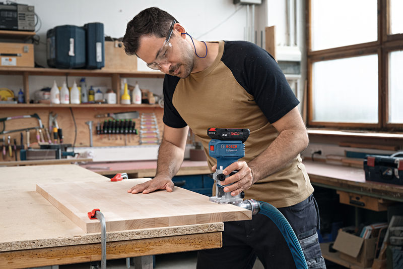 Eine Person mit Sicherheitsausrüstung formt mit einem handgeführten Elektrowerkzeug ein Holzbrett. Anwendungsbild