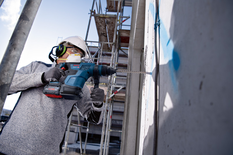Person mit Sicherheitsausrüstung bohrt auf einer Baustelle in eine Betonwand. Anwendungsbild
