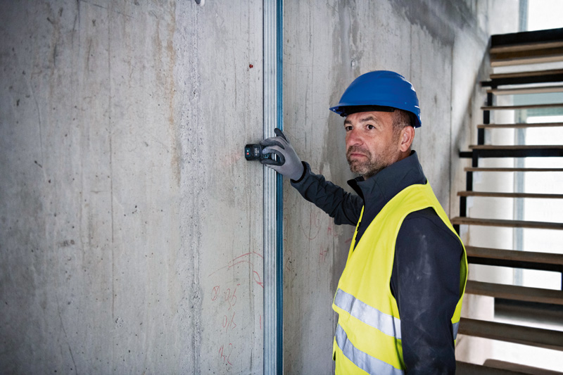 Eine Person mit Sicherheitsausrüstung verwendet ein Lasermessgerät an einer Betonwand. Anwendungsbild