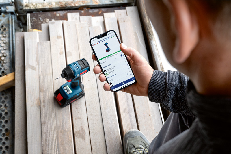 Eine Person prüft auf einem Smartphone neben einer Akku-Bohrmaschine die App-Funktionen eines Elektrowerkzeugs. Anwendungsbild