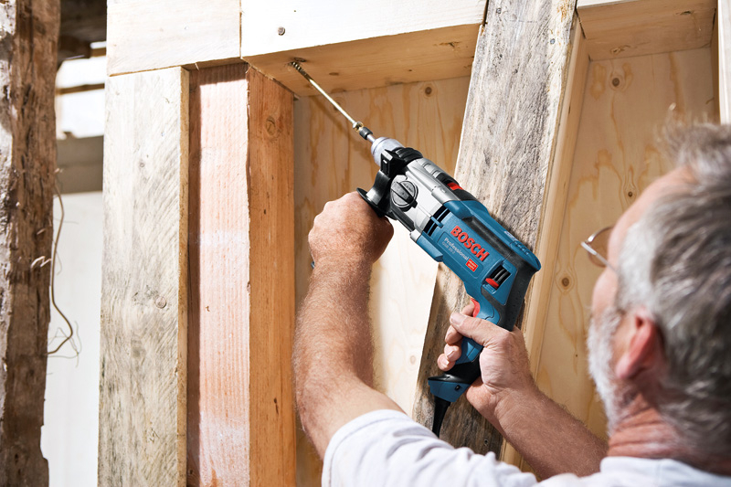 Eine Person bedient auf einer Baustelle eine Schlagbohrmaschine an einem Holzbalken. Anwendungsbild