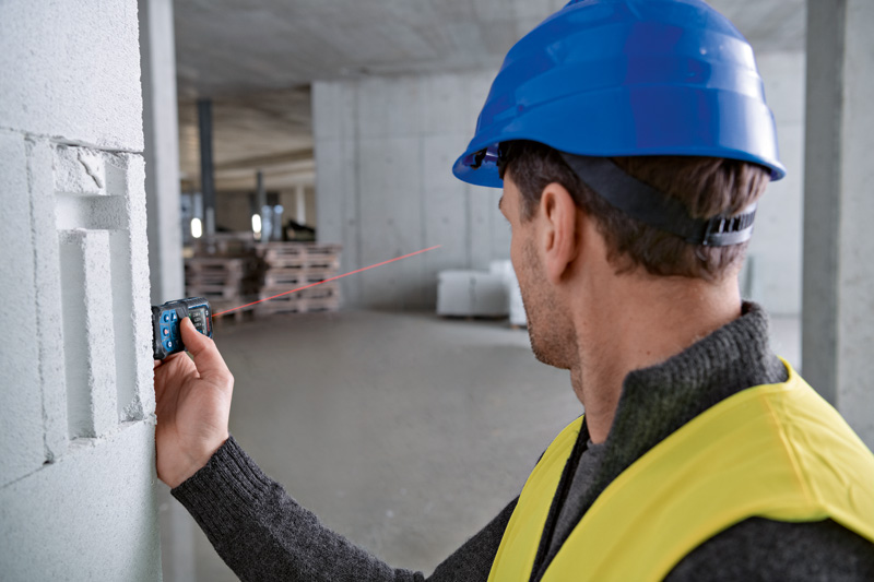 Eine Person mit Sicherheitsausrüstung verwendet ein Lasermessgerät, um auf einer Baustelle Entfernungsmessungen vorzunehmen. Anwendungsbild