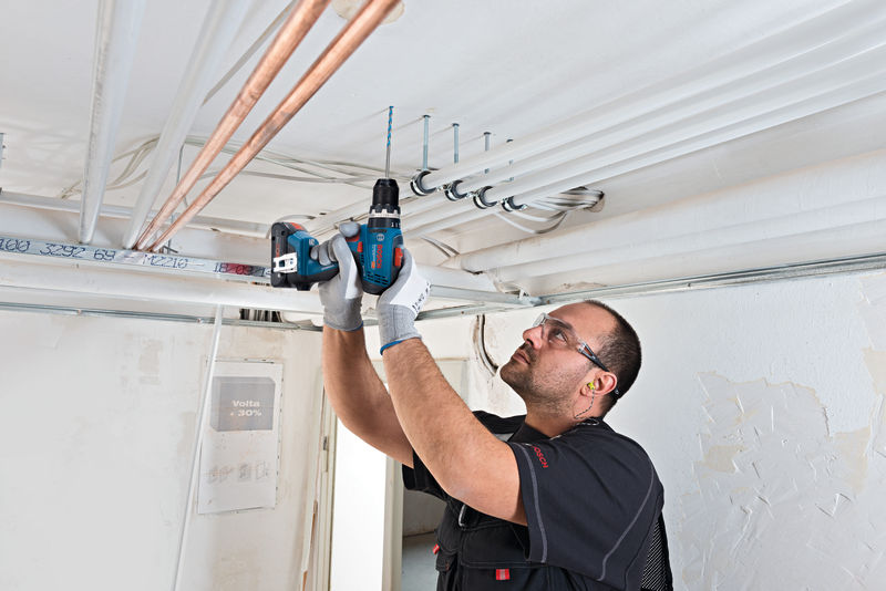 Eine Person mit Sicherheitsausrüstung bohrt in eine Decke, um elektrische Leitungen zu installieren. Anwendungsbild