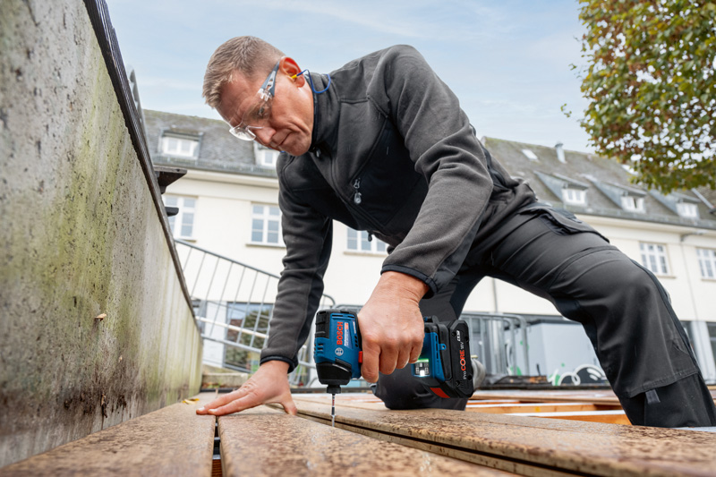 Eine Person mit Sicherheitsausrüstung bohrt in Holzbretter auf einem Außendeck. Anwendungsbild